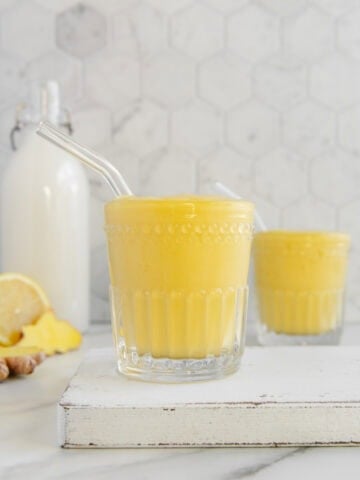 Two glasses of ginger smoothie with glass straws with a bottle of almond milk and sliced lemon and fresh ginger in the background on a white marble surface.