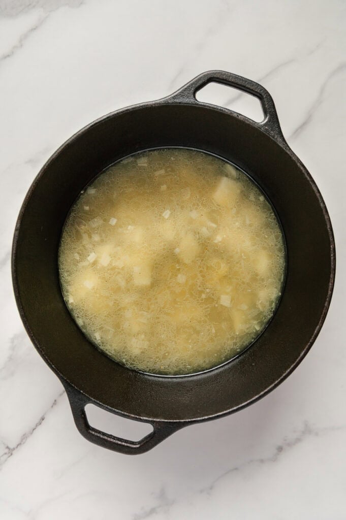 Boiled chopped russet potatoes in a dutch oven covered in water with a white marble background.