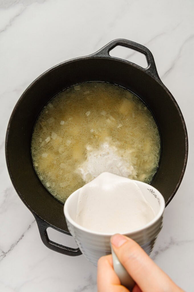 Vegan heavy cream being poured from a white measuring cup into a dutch oven with boiled russet potatoes.