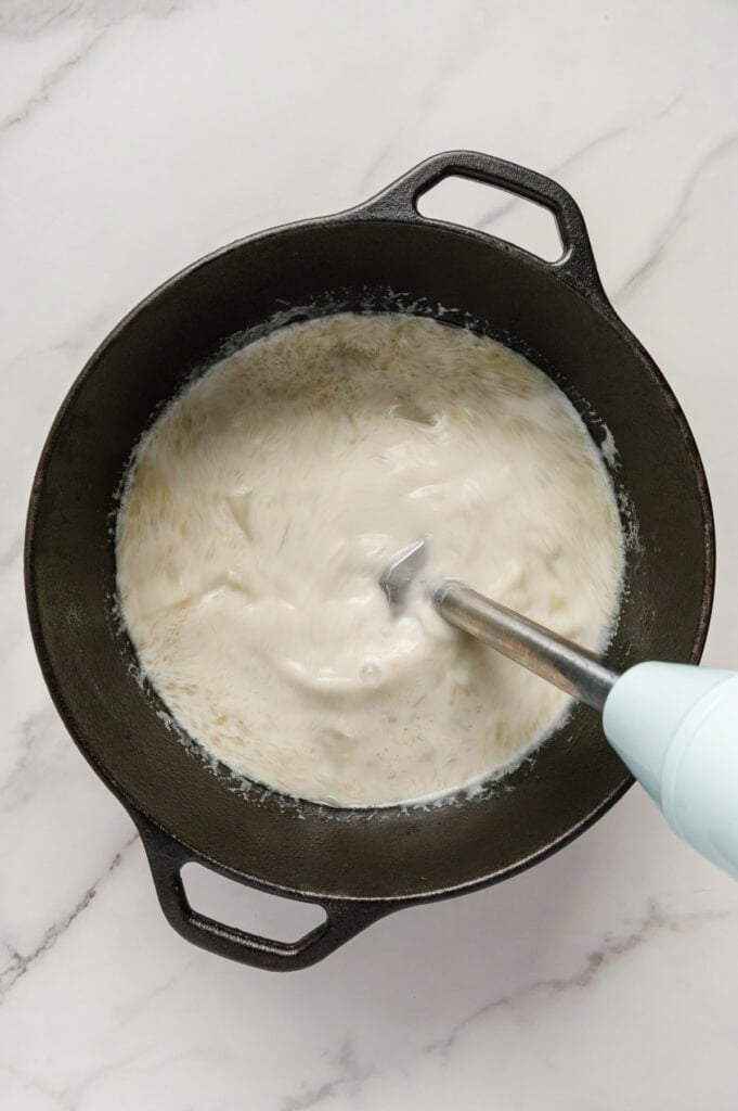 An immersion blender being used to blend vegan potato soup in a dutch oven.
