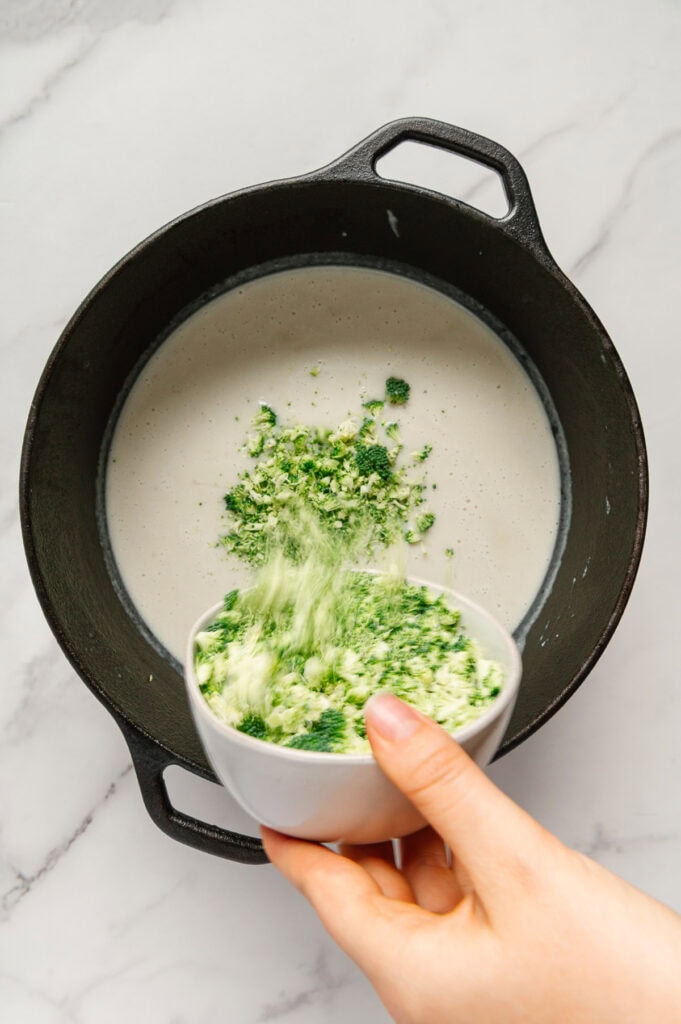 A hand pouring broccoli florets from a bowl into vegan potato soup in a dutch oven.