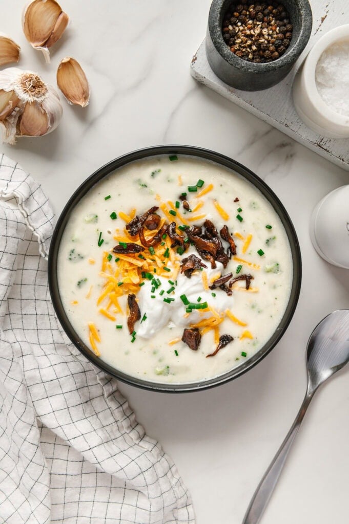 Vegan potato soup garnished with vegan shredded cheddar cheese, vegan sour cream, mushroom bacon and chives with a white marble background.