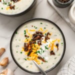 Vegan potato soup garnished with vegan shredded cheddar cheese, vegan sour cream, mushroom bacon and chives with a white marble background.
