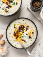 Vegan potato soup garnished with vegan shredded cheddar cheese, vegan sour cream, mushroom bacon and chives with a white marble background.