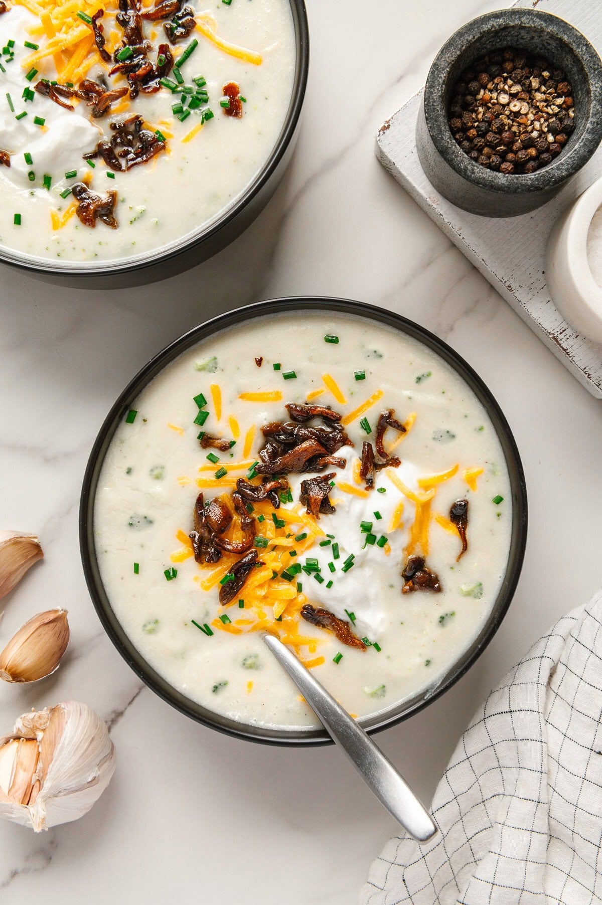 Vegan potato soup garnished with vegan shredded cheddar cheese, vegan sour cream, mushroom bacon and chives with a white marble background.