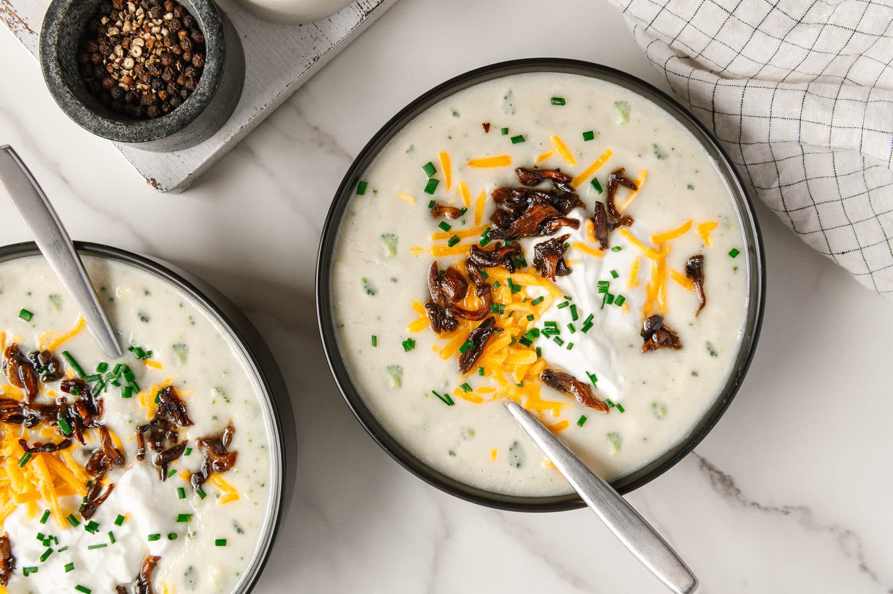 Two bowls of vegan potato soup garnished with vegan shredded cheddar cheese, vegan sour cream, mushroom bacon and chives with a white marble background.