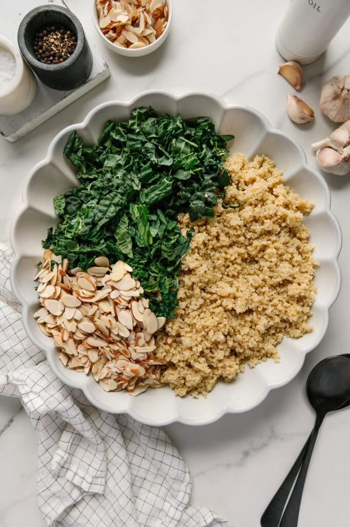 Kale, quinoa and sliced almonds in a white serving bowl next to fresh garlic, salt and pepper with a white marble background.