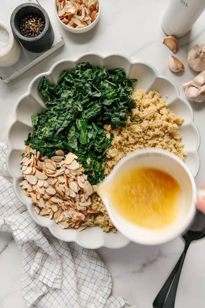 Pouring a lemon garlic dressing over kale, quinoa and sliced almonds in a white serving bowl.