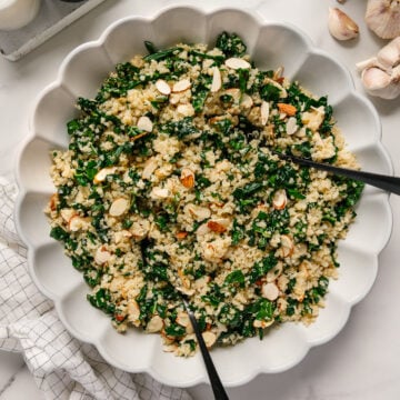 Kale quinoa salad with sliced almonds in a white serving bowl with black salad tongs all on a white background with garlic off to the side and salt and pepper