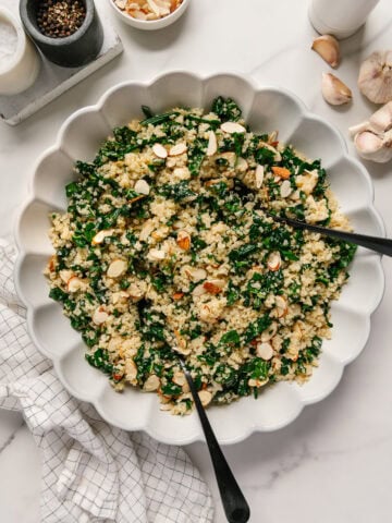 Kale quinoa salad with sliced almonds in a white serving bowl with black salad tongs all on a white background with garlic off to the side and salt and pepper