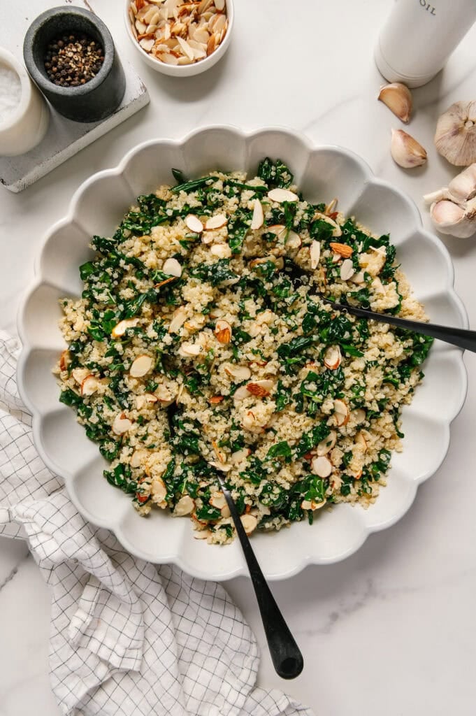 Kale quinoa salad in a white serving bowl next to fresh garlic, salt and pepper with a white marble background.