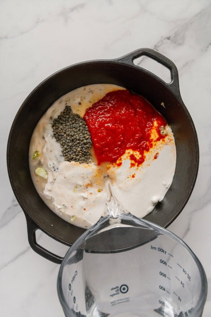 Marinara sauce, vegan heavy cream and lentils added to cabbage soup ingredients while water is being poured into a dutch oven with a white marble background.