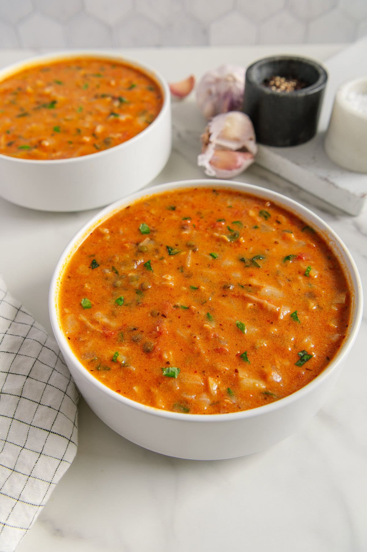 Vegan cabbage soup in two white bowls with fresh garlic and dry lentils in the background with a white background.