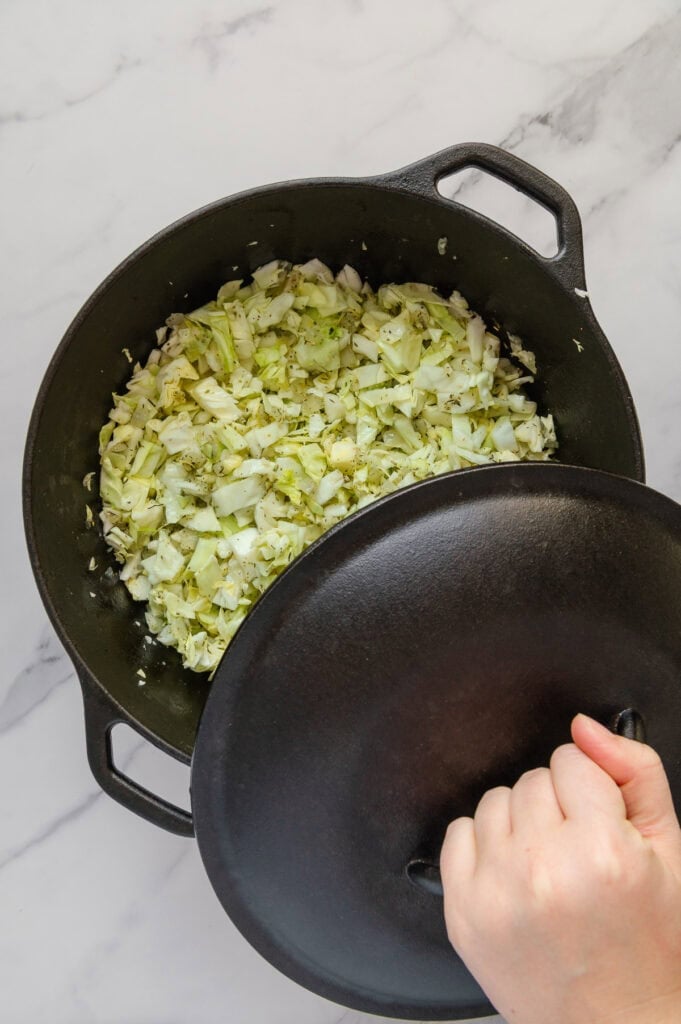 A hand putting a lid over a dutch oven full of cabbage with a white marble background..