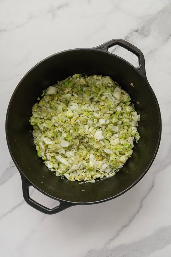 Green cabbage wilted down in a dutch oven with other cabbage soup ingredients with a white marble background.