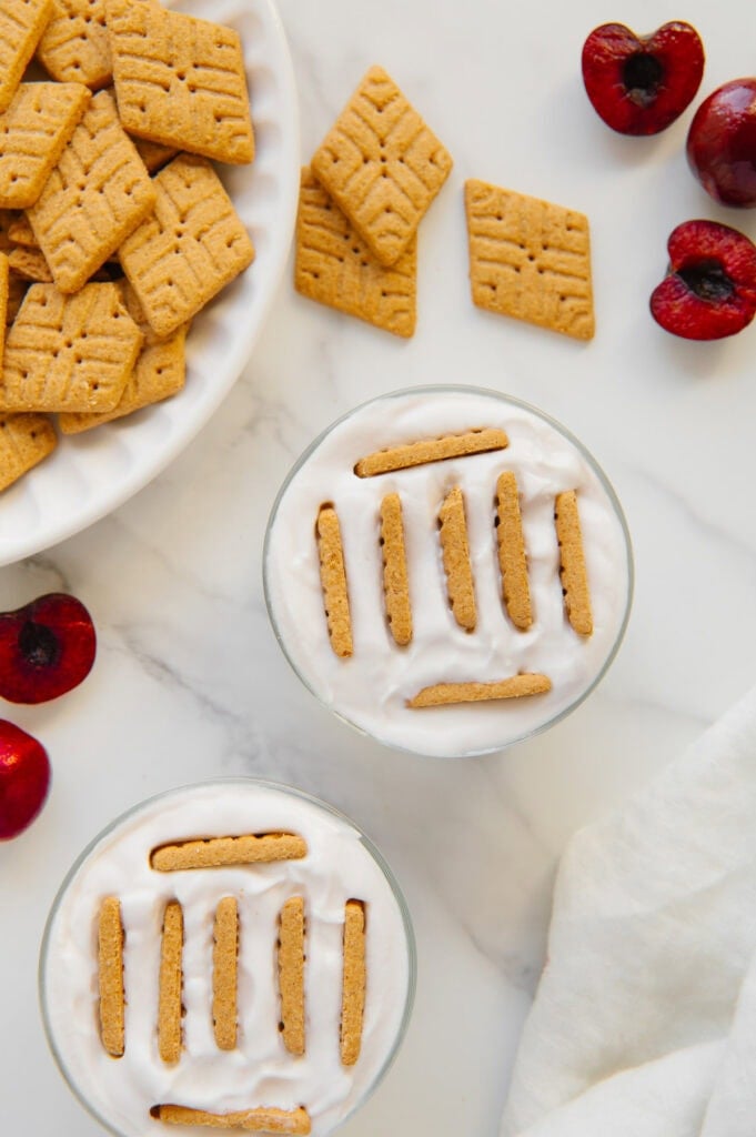 Simple Mills cookies placed vertically into two glasses of yogurt with a white marble background.