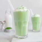 Two glasses of matcha smoothie topped with whipped cream and sifted matcha with matcha and a bottle of milk in the background.