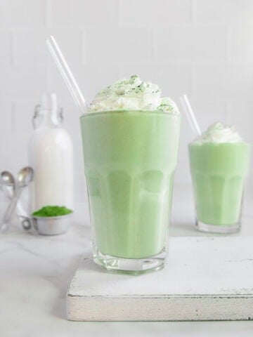 Two glasses of matcha smoothie topped with whipped cream and sifted matcha with matcha and a bottle of milk in the background.