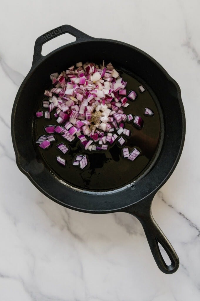 Diced red onions and garlic in a cast iron skillet with a white marble background.