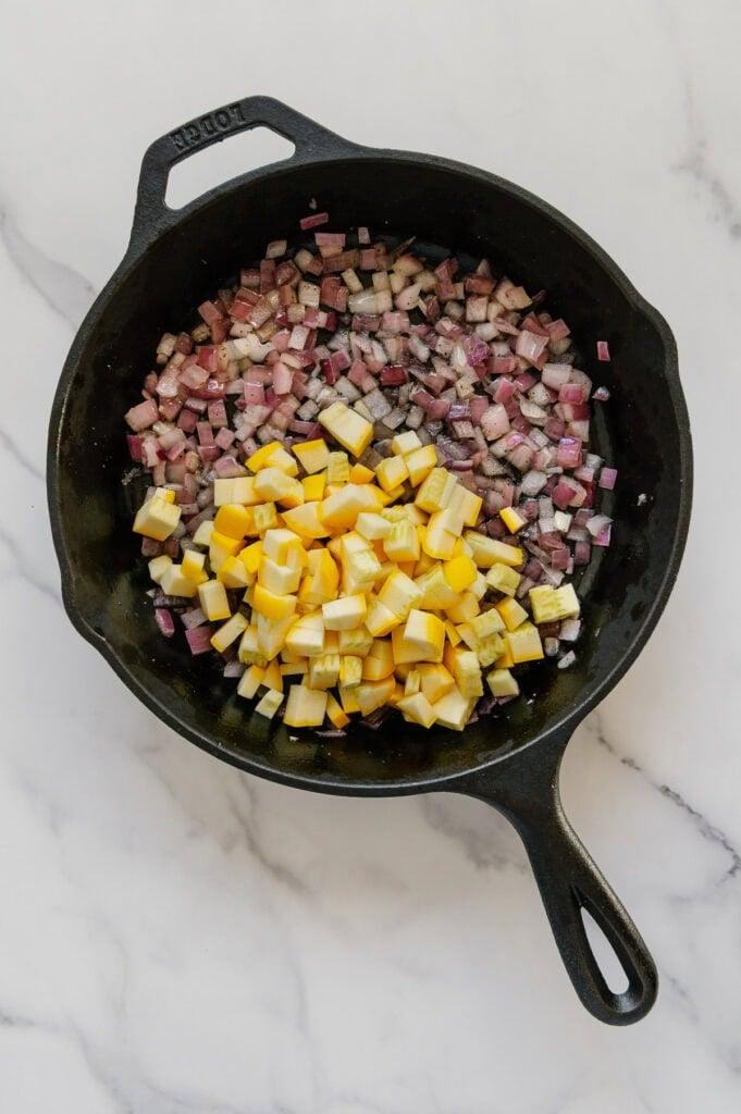 Diced yellow squash added to a cast iron skillet containing sautéed red onions and garlic.