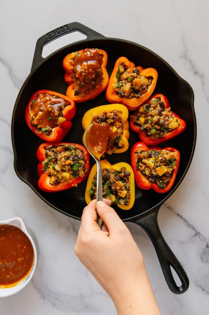 A hand spooning homemade enchilada sauce over stuffed bell peppers in a cast iron skillet.