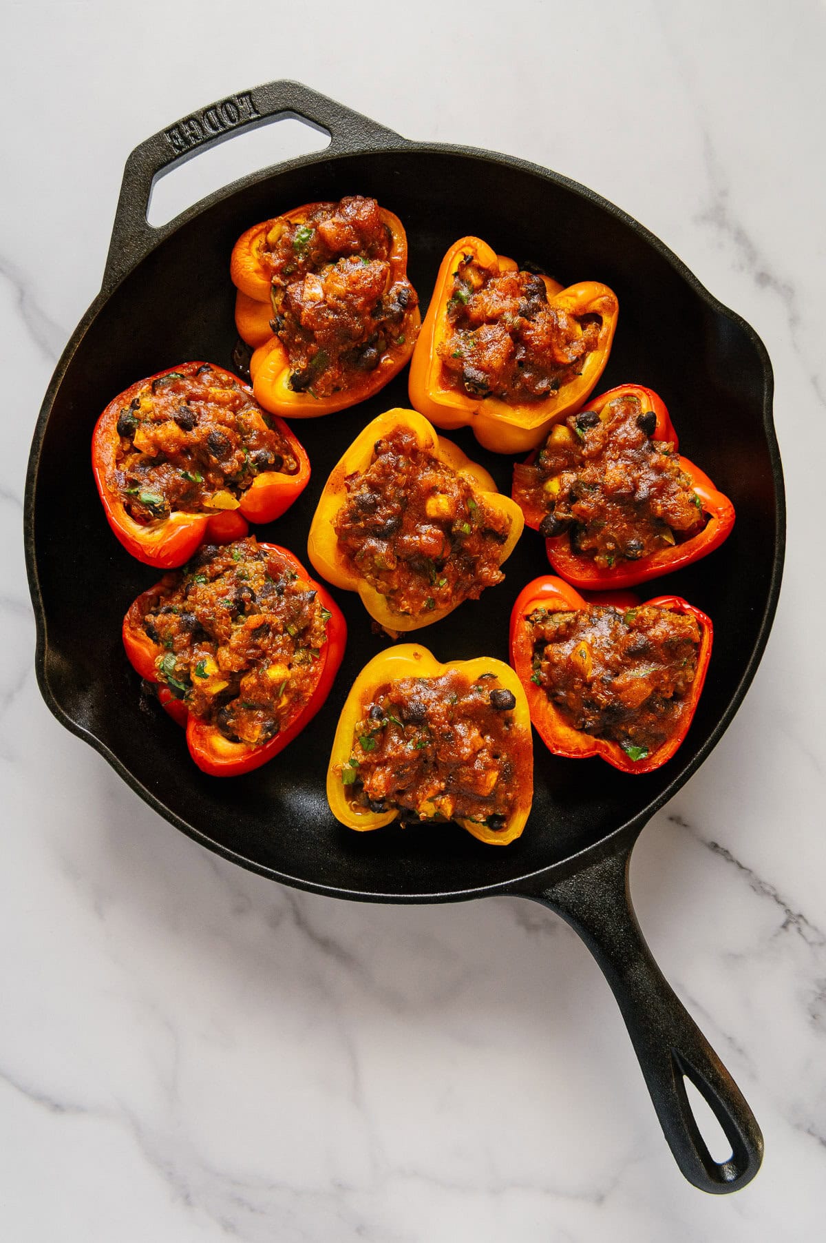 Eight roasted enchilada stuffed bell peppers in a cast iron skillet with a white marble background.