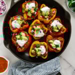 Vegan enchilada stuffed bell peppers topped with vegan sour cream, diced avocado, fresh cilantro and diced red onion in a cast iron skillet with a white marble background.