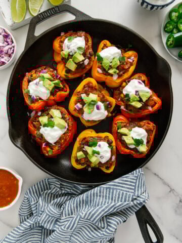 Vegan enchilada stuffed bell peppers topped with vegan sour cream, diced avocado, fresh cilantro and diced red onion in a cast iron skillet with a white marble background.