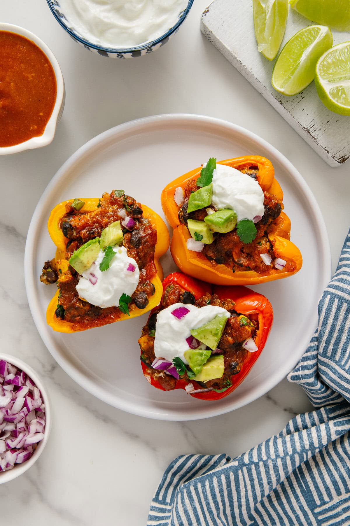Three enchilada stuffed bell peppers (red, yellow and orange) arranged on a white plate topped with sliced avocado, fresh cilantro and vegan sour cream.