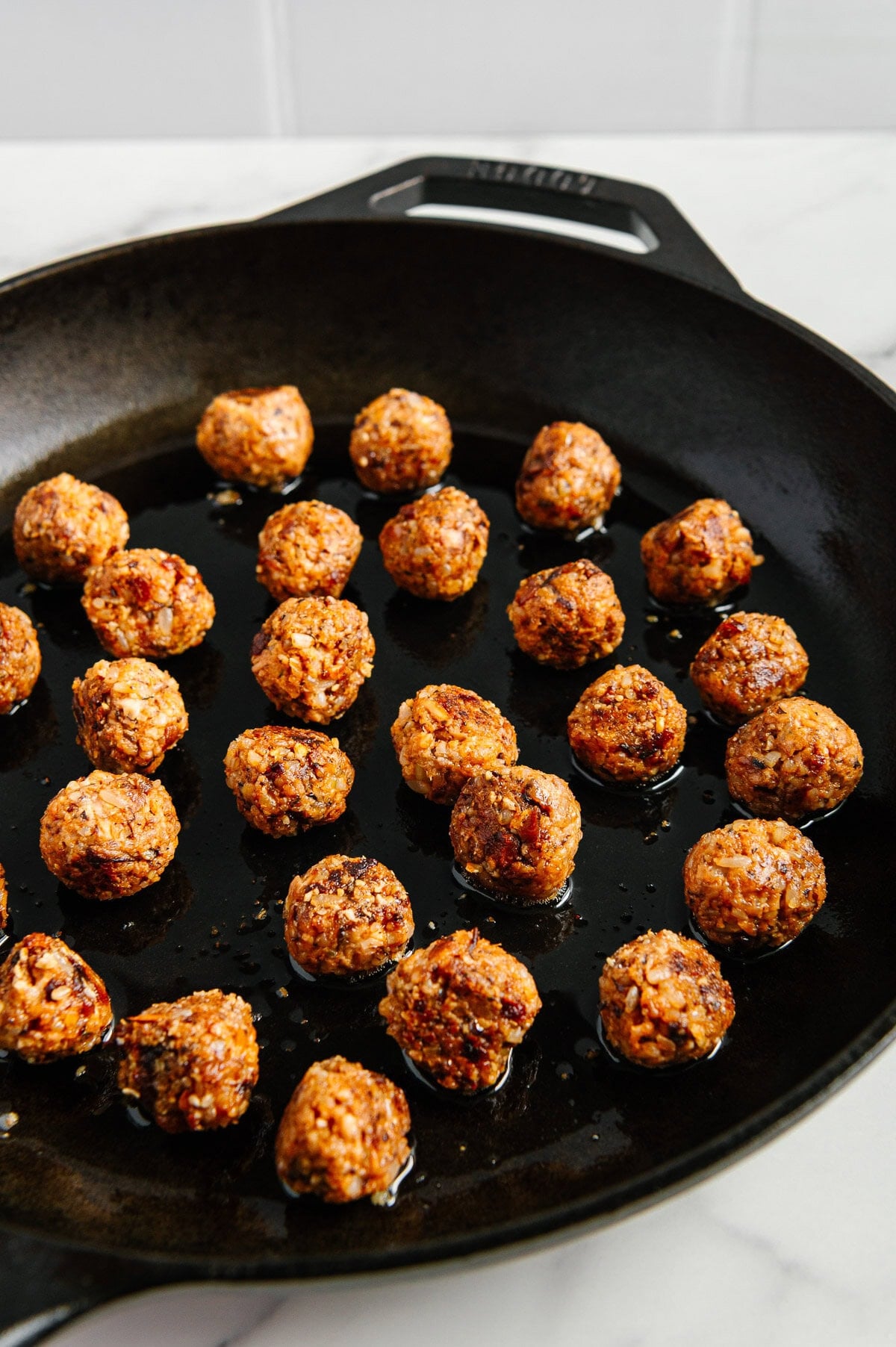 A black cast iron skillet with several small, browned vegan meatballs-perfect for vegan Italian wedding soup-cooking in oil, arranged evenly across the pan on a white marble surface.