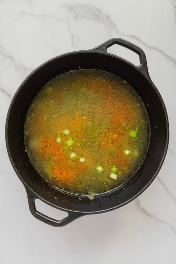 A black cast iron pot filled with vegan Italian wedding soup-broth, chopped carrots, green onions, and herbs-sits on a white marble surface.