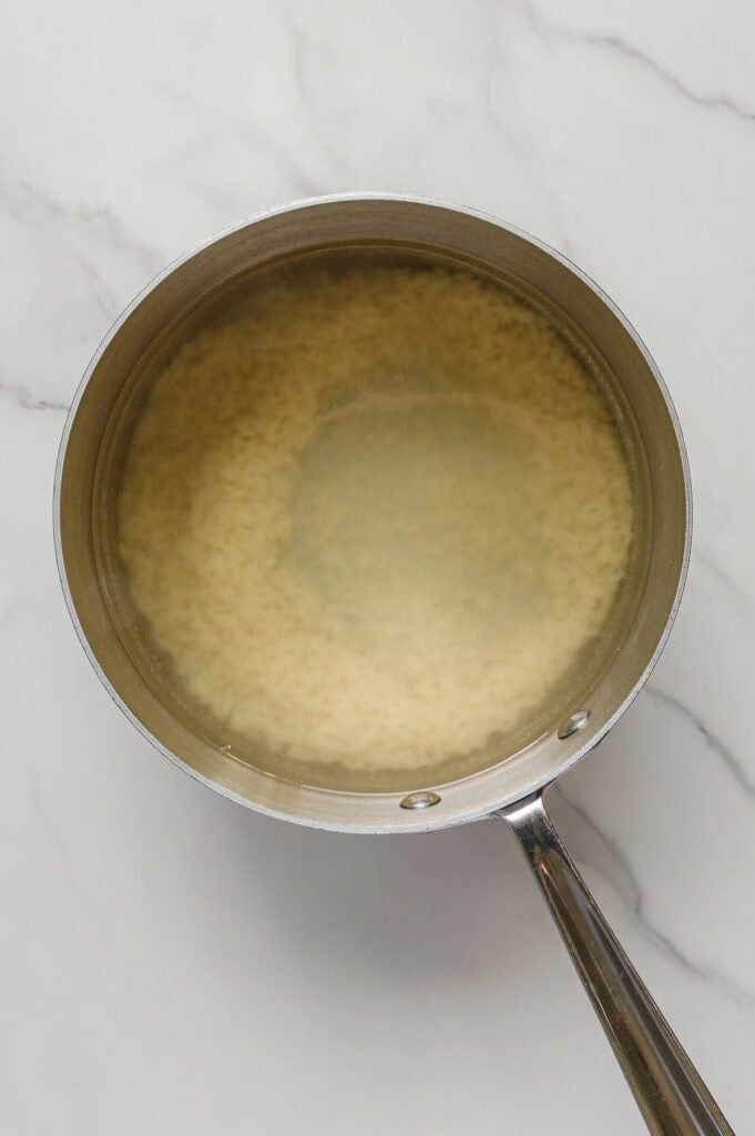 A stainless steel saucepan filled with water and uncooked rice, perfect for preparing vegan Italian wedding soup, sits on a white marble surface.