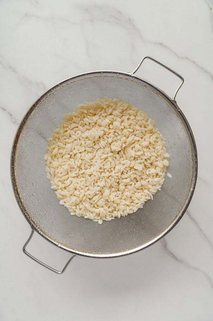 A metal strainer filled with cooked orzo pasta sits on a white marble surface, ready to be added to a comforting vegan Italian wedding soup.