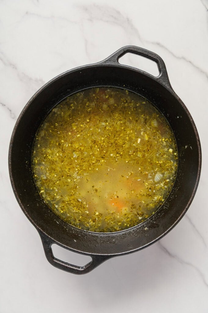 A black pot filled with vegan Italian wedding soup, its yellow-green broth brimming with finely chopped vegetables, sits on a white marble surface.