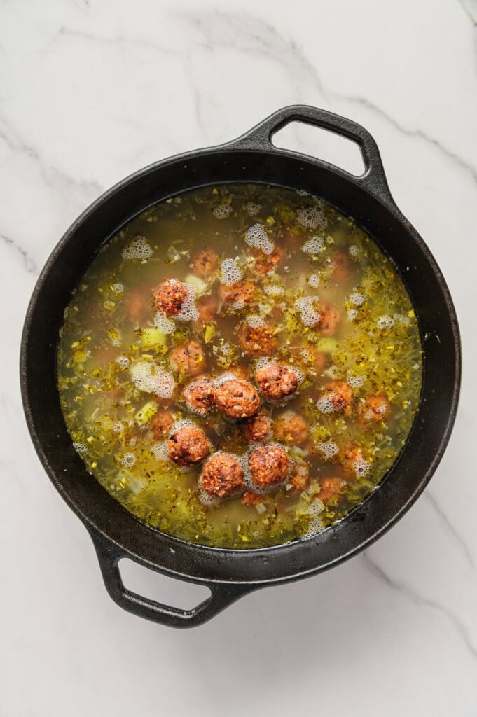 A black pot filled with vegan Italian wedding soup-featuring plant-based meatballs, chopped vegetables, and savory broth-sits on a white marble surface.