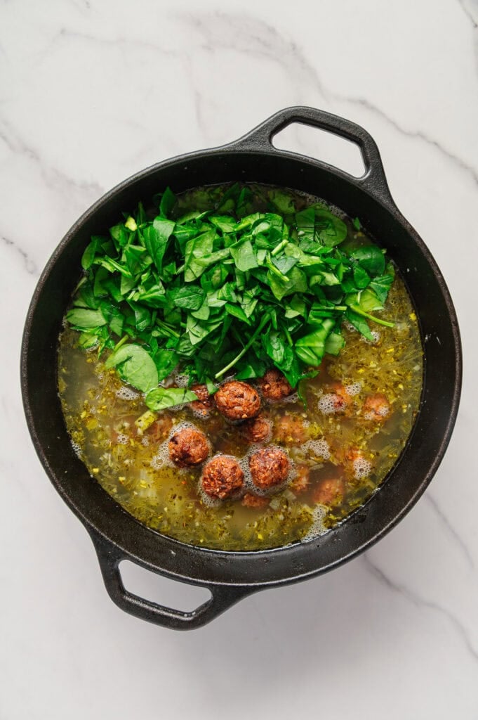 A black pot on a marble surface contains vegan Italian wedding soup with plant-based meatballs, savory broth, and a pile of fresh chopped spinach on top, ready to be mixed in.