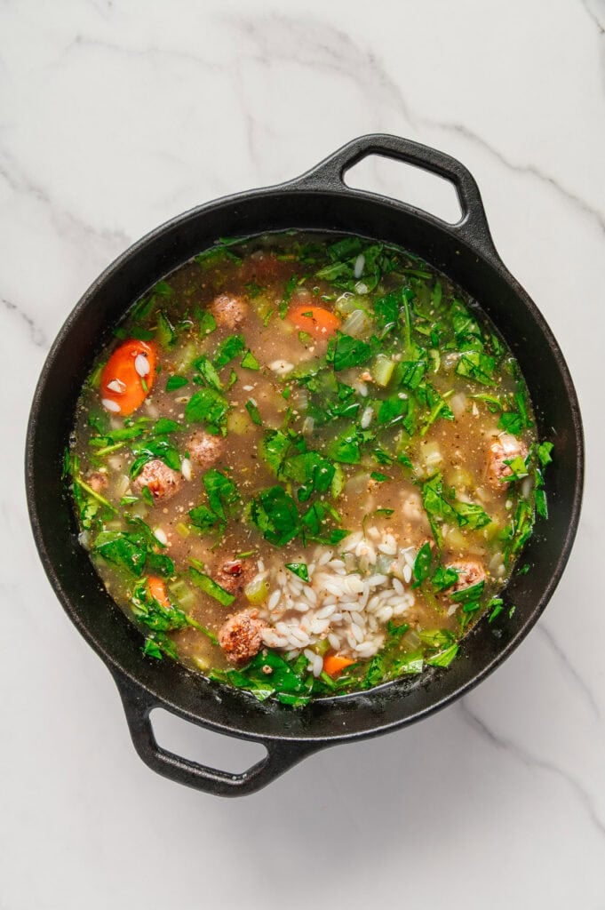 A black pot filled with vegan Italian wedding soup featuring rice, chopped spinach, carrots, celery, and herbs sits on a white marble surface.
