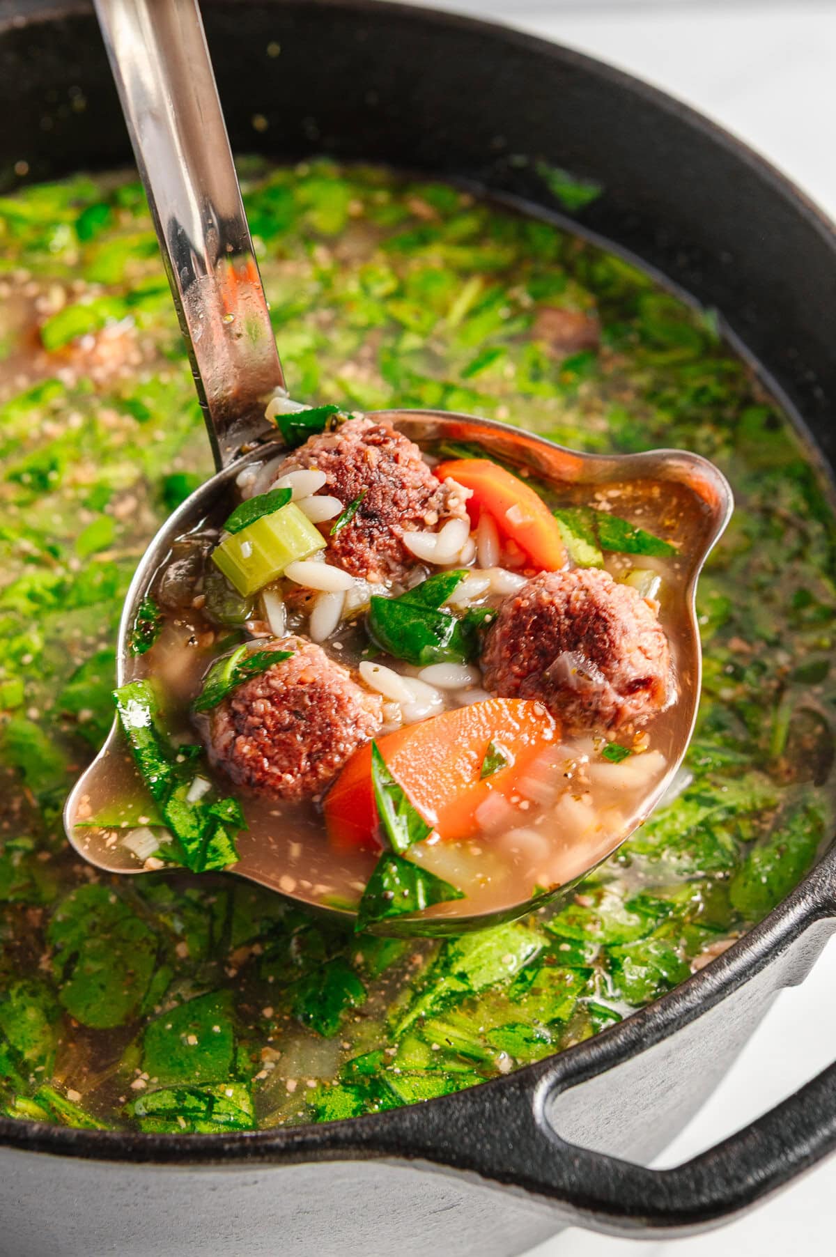 A ladle lifts vegan Italian wedding soup with rice, carrots, celery, and spinach from a black pot filled with the same colorful, hearty soup.
