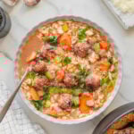 A bowl of vegan Italian wedding soup with meatballs, orzo pasta, carrots, celery, greens, and broth sits on a marble surface, surrounded by garlic cloves, grated cheese, and a napkin. A spoon rests in the bowl.