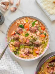A bowl of vegan Italian wedding soup with meatballs, orzo pasta, carrots, celery, greens, and broth sits on a marble surface, surrounded by garlic cloves, grated cheese, and a napkin. A spoon rests in the bowl.