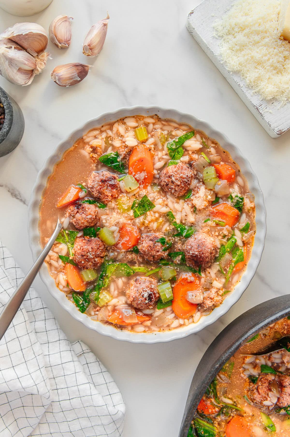 A bowl of vegan Italian wedding soup with meatballs, orzo pasta, carrots, celery, greens, and broth sits on a marble surface, surrounded by garlic cloves, grated vegan cheese, and a napkin. A spoon rests in the bowl.