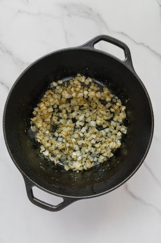 A black cast iron pot containing sautéed diced onions and seasoning for vegan Italian wedding soup sits on a white marble surface.