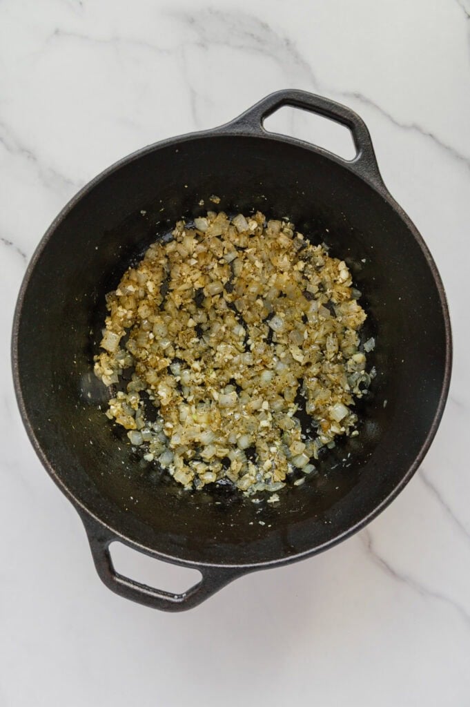 Chopped onions and garlic sautéing in a black cast iron pot on a white marble countertop, forming the flavorful base for a vegan Italian wedding soup.