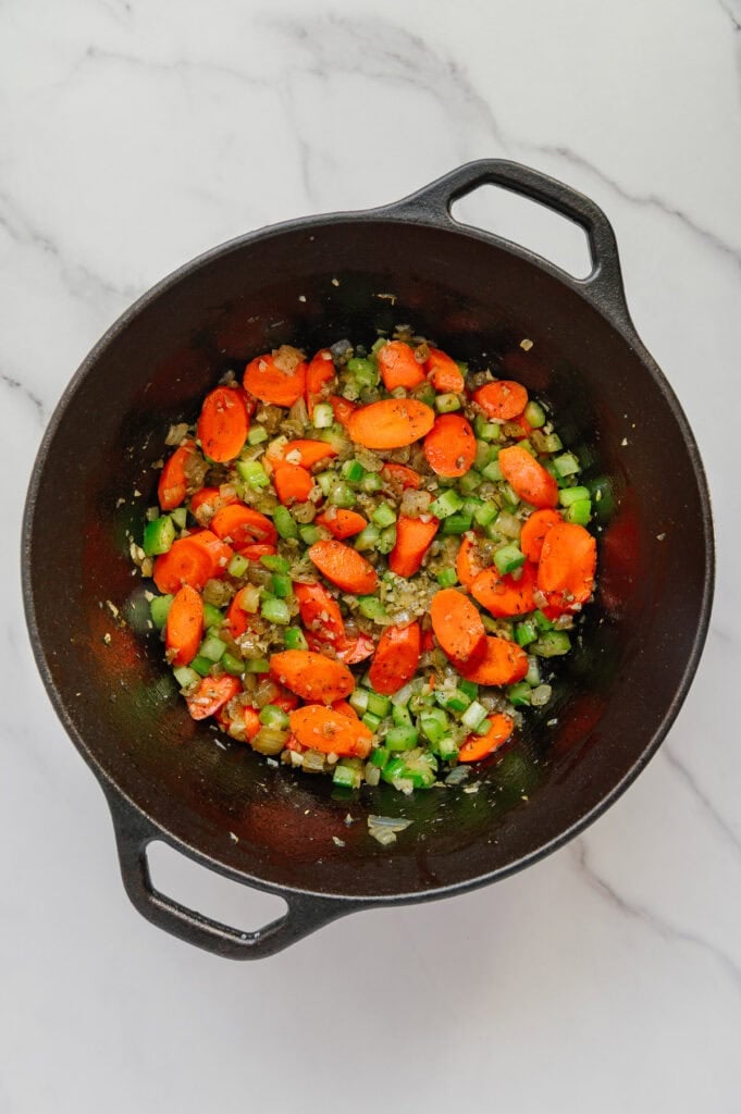 A black pot on a marble surface contains chopped carrots, green bell peppers, celery, and onions being sautéed together as the hearty base for a vegan Italian wedding soup.