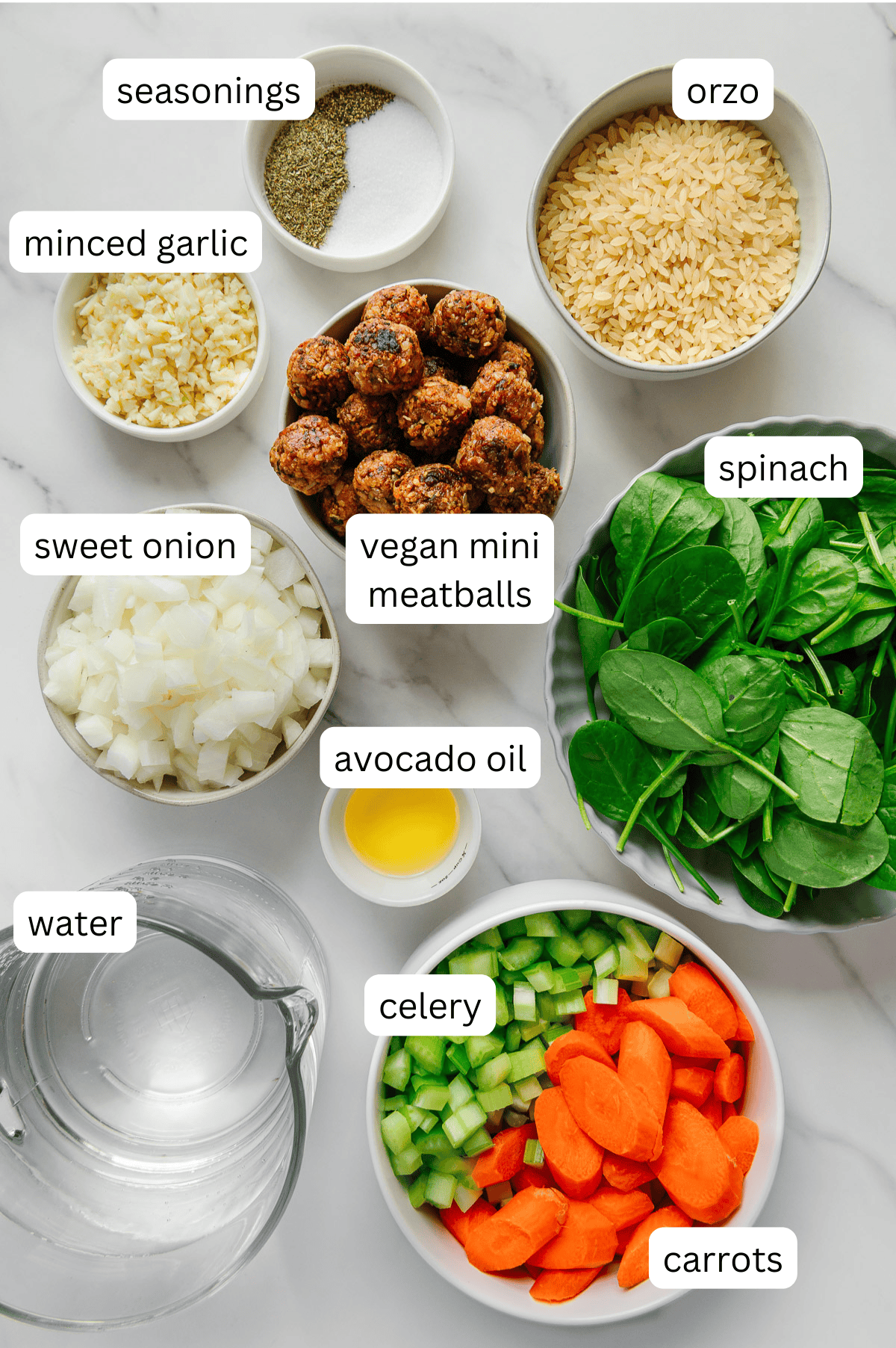 Overhead view of labeled ingredients for vegan Italian wedding soup: seasonings, orzo, minced garlic, vegan mini meatballs, chopped sweet onion, spinach leaves, avocado oil, water, chopped celery, and sliced carrots.