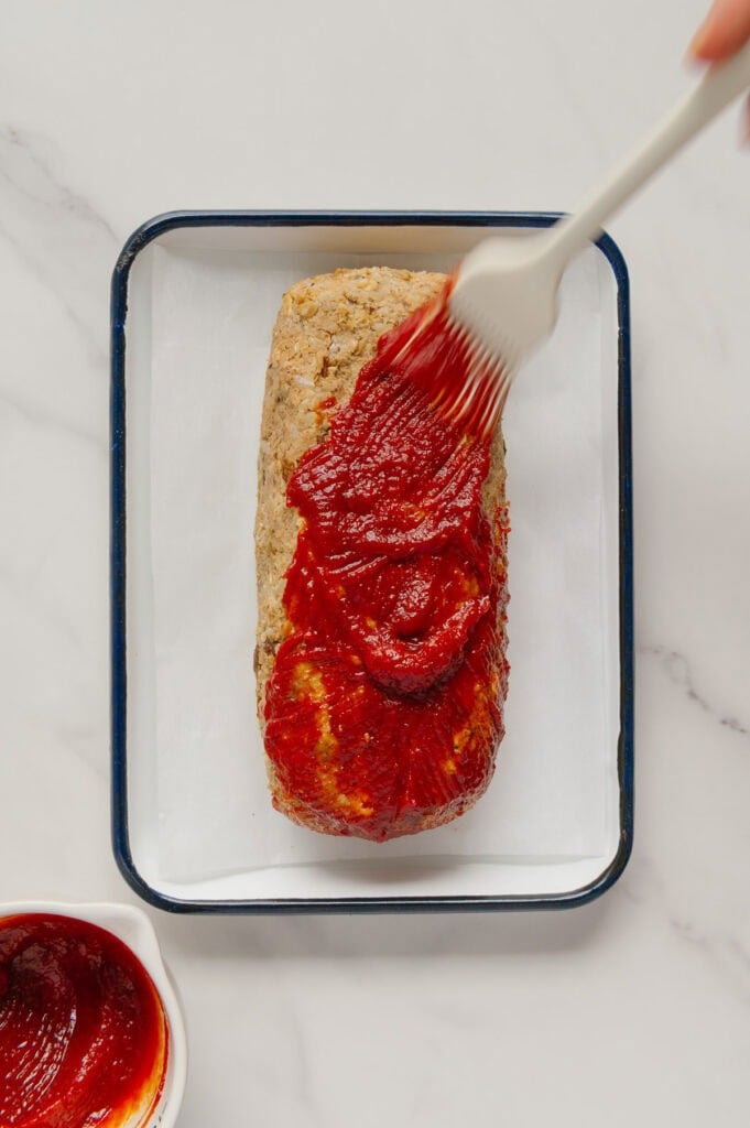 A hand uses a brush to spread red sauce over an uncooked loaf on a parchment-lined baking pan. A small bowl with more sauce sits nearby on a white marble surface.