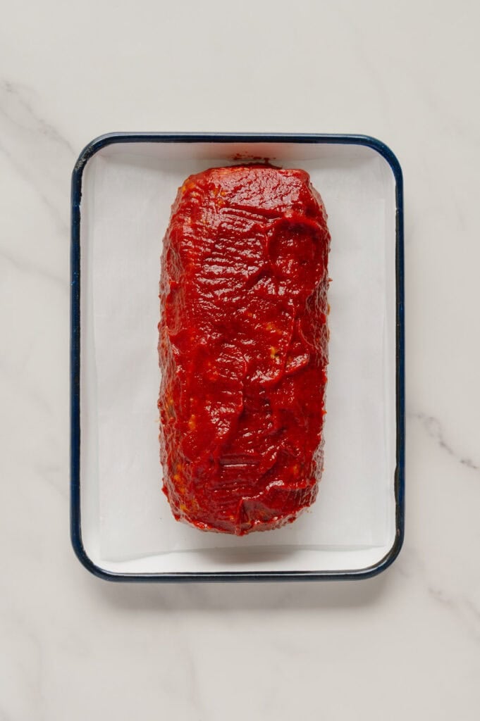A rectangular, uncooked vegan meatloaf covered in a red sauce sits on parchment paper in a black-edged baking tray on a white marble surface.