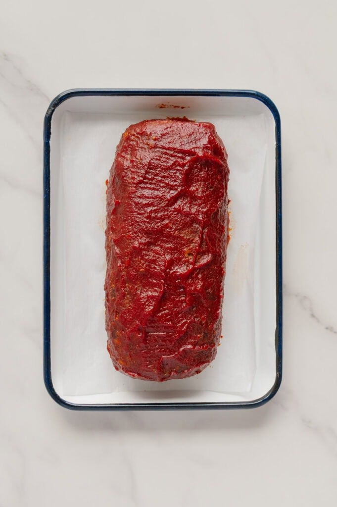 A rectangular, cooked vegan meatloaf coated with a red sauce sits on a parchment-lined baking tray, set on a light-colored marble surface.
