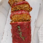 A loaf of vegan meatloaf topped with tomato sauce, partially sliced, is displayed on a white cutting board. A sprig of fresh thyme garnishes the top. The background includes a napkin and marble surface.