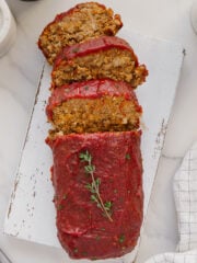 A loaf of vegan meatloaf topped with tomato sauce, partially sliced, is displayed on a white cutting board. A sprig of fresh thyme garnishes the top. The background includes a napkin and marble surface.
